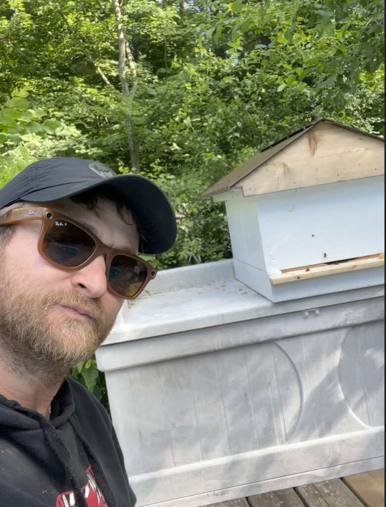 A beekeeper in a cap and sunglasses standing beside a white beehive with a gabled roof, in a wooded backyard.